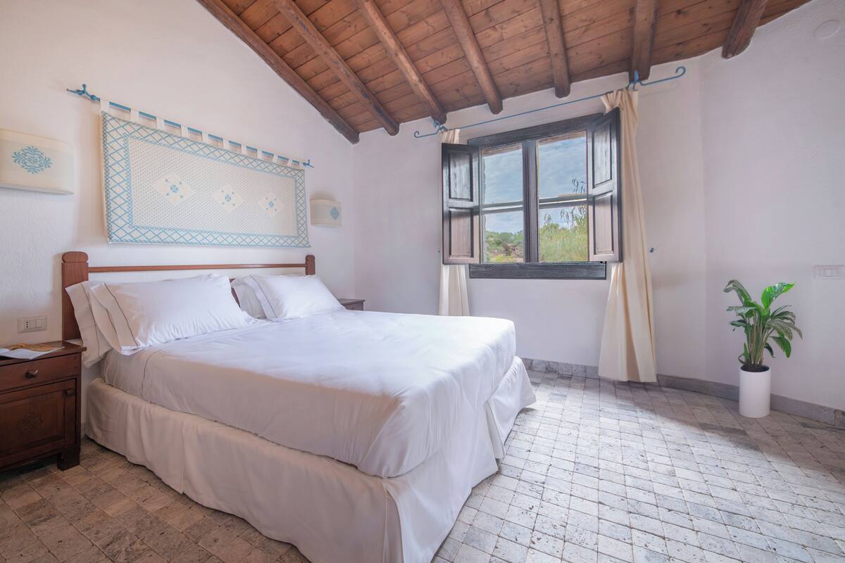 A bedroom with a wooden ceiling, a bed with white bedding, a nightstand, a window with curtains, and a potted plant.