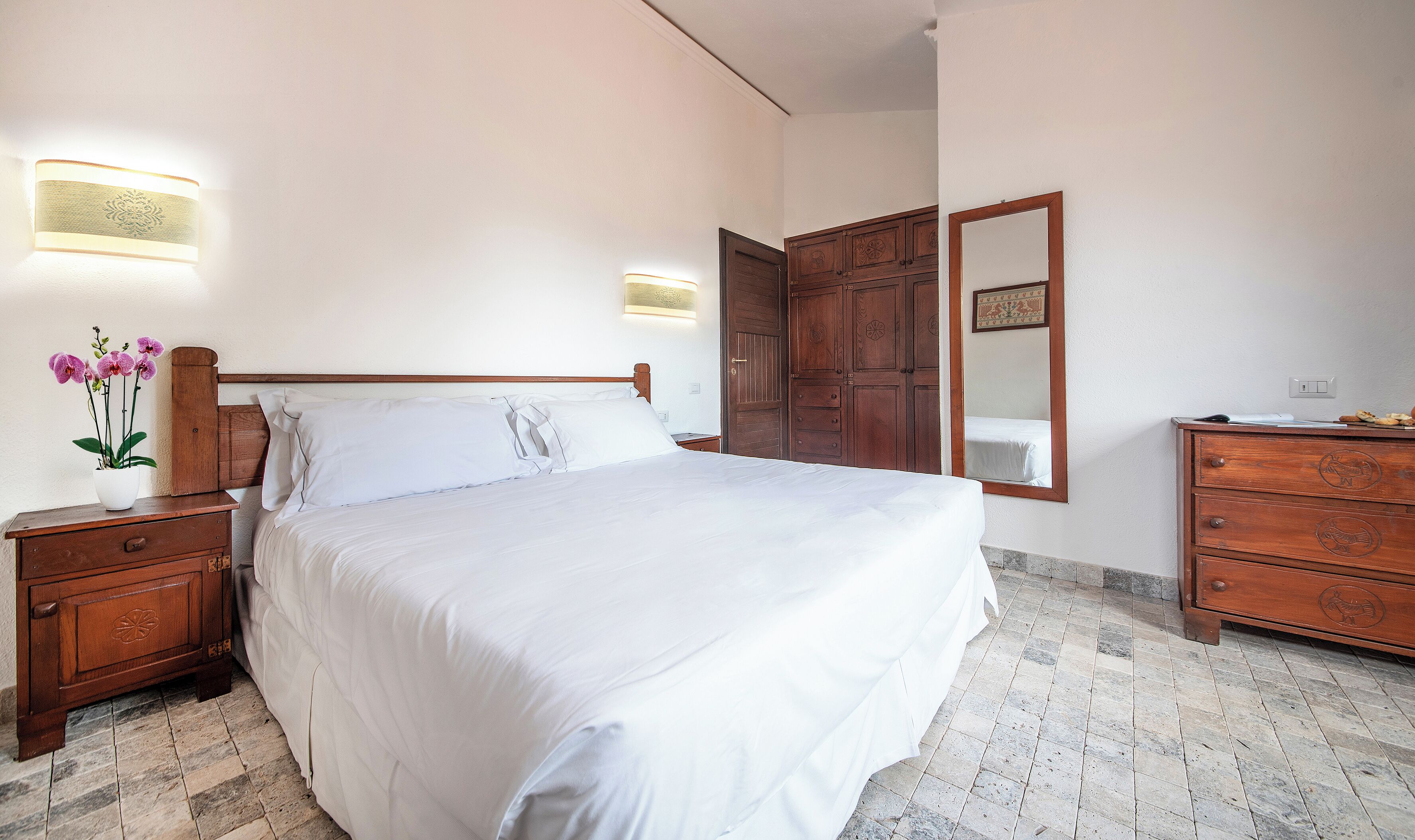 A bedroom with a large bed, wooden nightstands, a dresser, a mirror, and a wall-mounted light fixture.