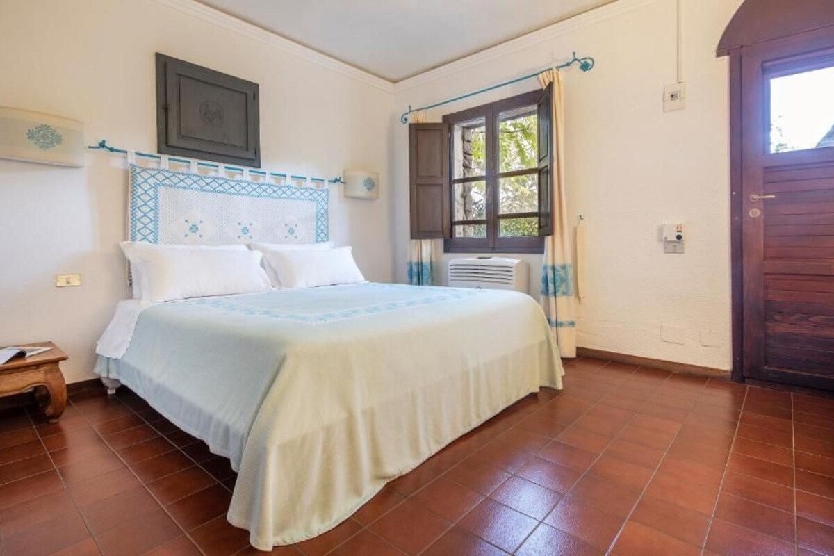 Un dormitorio con cama, mesita de noche, ventana con cortinas y puerta.