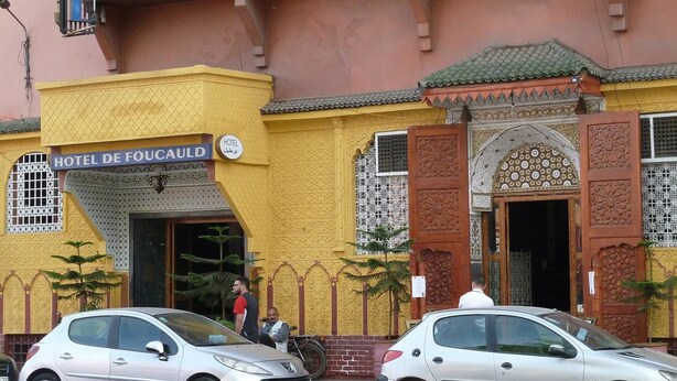 Fachada de hotel con detalles arquitectónicos intrincados, un cartel que dice "HOTEL DE FOCAULD" y dos coches estacionados.
