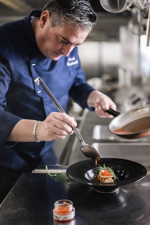 A chef in a blue uniform preparing a dish on a black plate. - Hotel Ladinia (Badia)