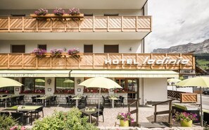 A hotel with a restaurant and snack bar, outdoor seating with umbrellas, and flower boxes on the balcony. - Hotel Ladinia (Badia)