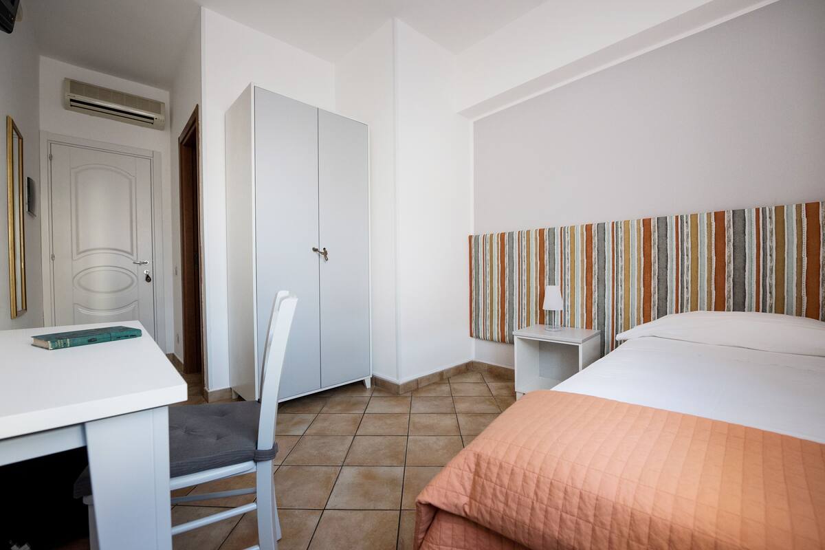 Une chambre d’hôtel avec un lit, un bureau, une chaise, une armoire et un rideau à rayures.