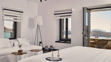 A modern bedroom with a large bed, two bedside tables with decorative items, a white sofa, and a view of the sea through a glass door.