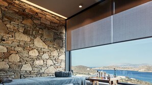 Una habitación con pared de piedra, un ventanal con vistas al mar, una cama con una toalla azul y una mesita con artículos de aseo.