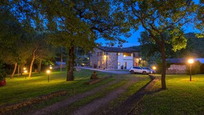 Une maison en pierre avec une allée pavée et une pelouse, entourée d’arbres, et une voiture blanche garée devant.
