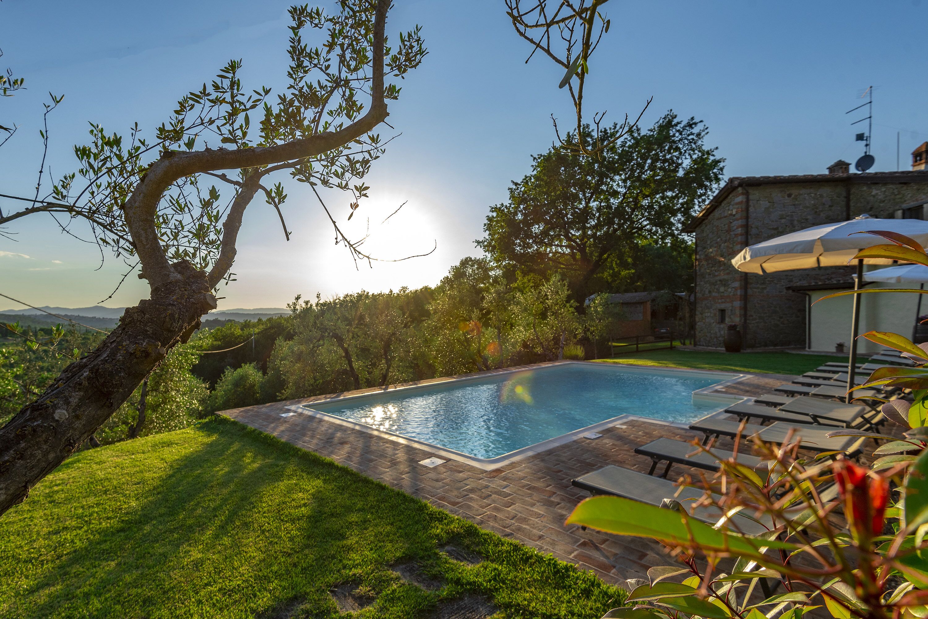 A swimming pool with lounge chairs and an umbrella, surrounded by greenery and a stone building.