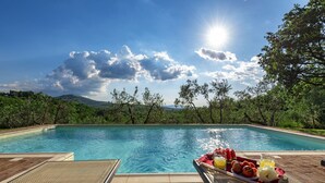 Une piscine, avec un plateau de petit-déjeuner composé de fruits et de jus, entourée d’arbres et sous un ciel dégagé.