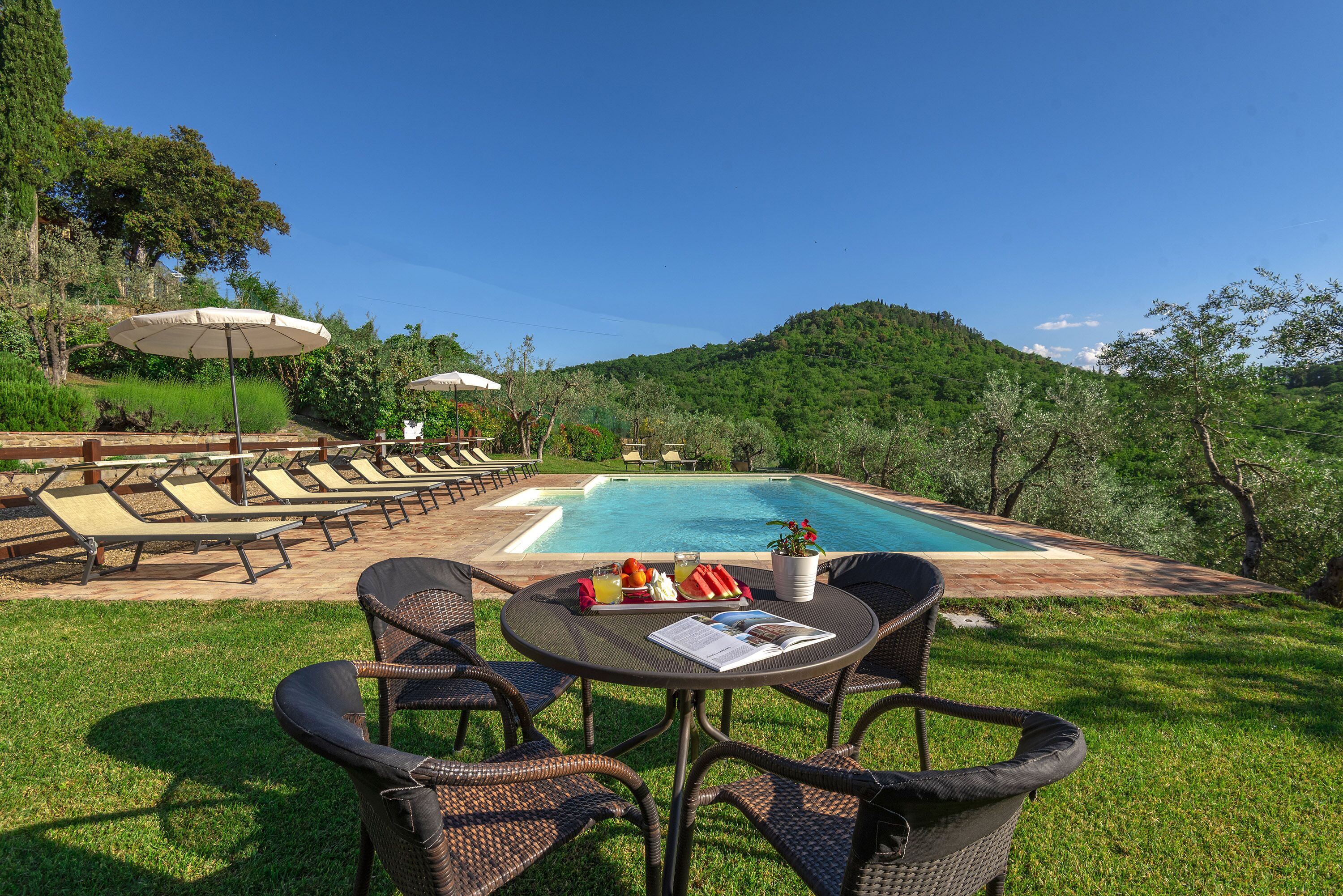 Zona de piscina con sillones, una mesa con un libro y fruta, y vista a la montaña.