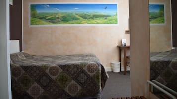 Une chambre avec deux lits, un tableau représentant un paysage accroché au mur, une petite table avec une lampe et une poubelle.