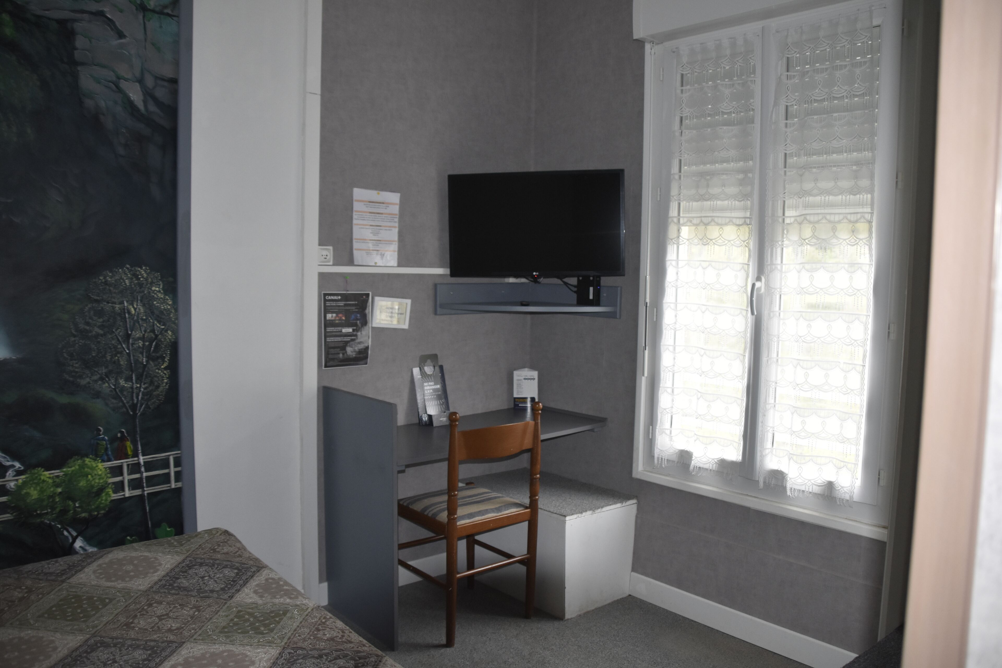 Une pièce avec un téléviseur fixé au mur, une chaise en bois, un petit bureau et une fenêtre ornée de rideaux légers.