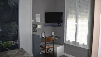 Une pièce avec un téléviseur fixé au mur, une chaise en bois, un petit bureau et une fenêtre ornée de rideaux légers.
