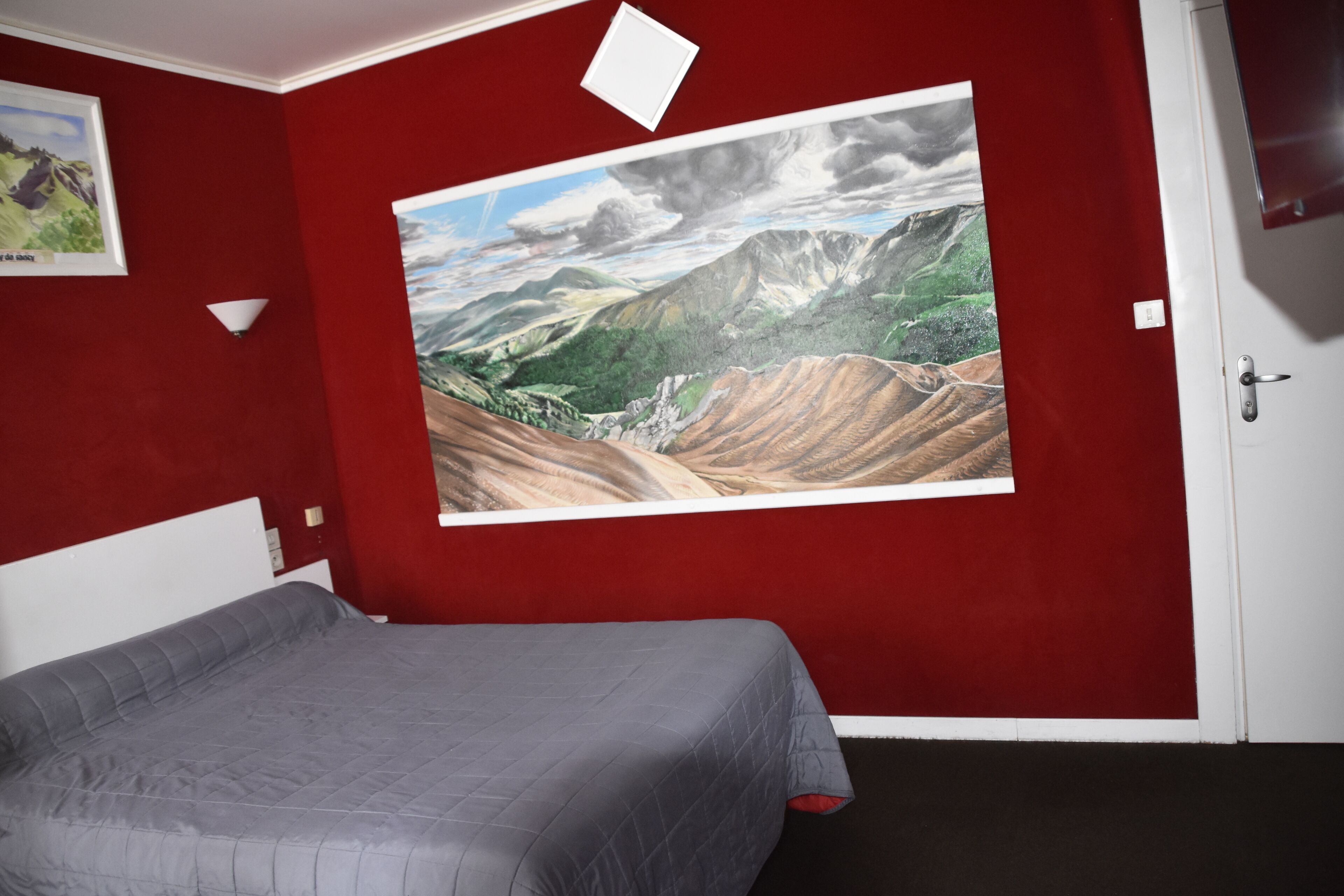 A room with a large landscape painting on the wall, a bed with a grey duvet, and a white door.