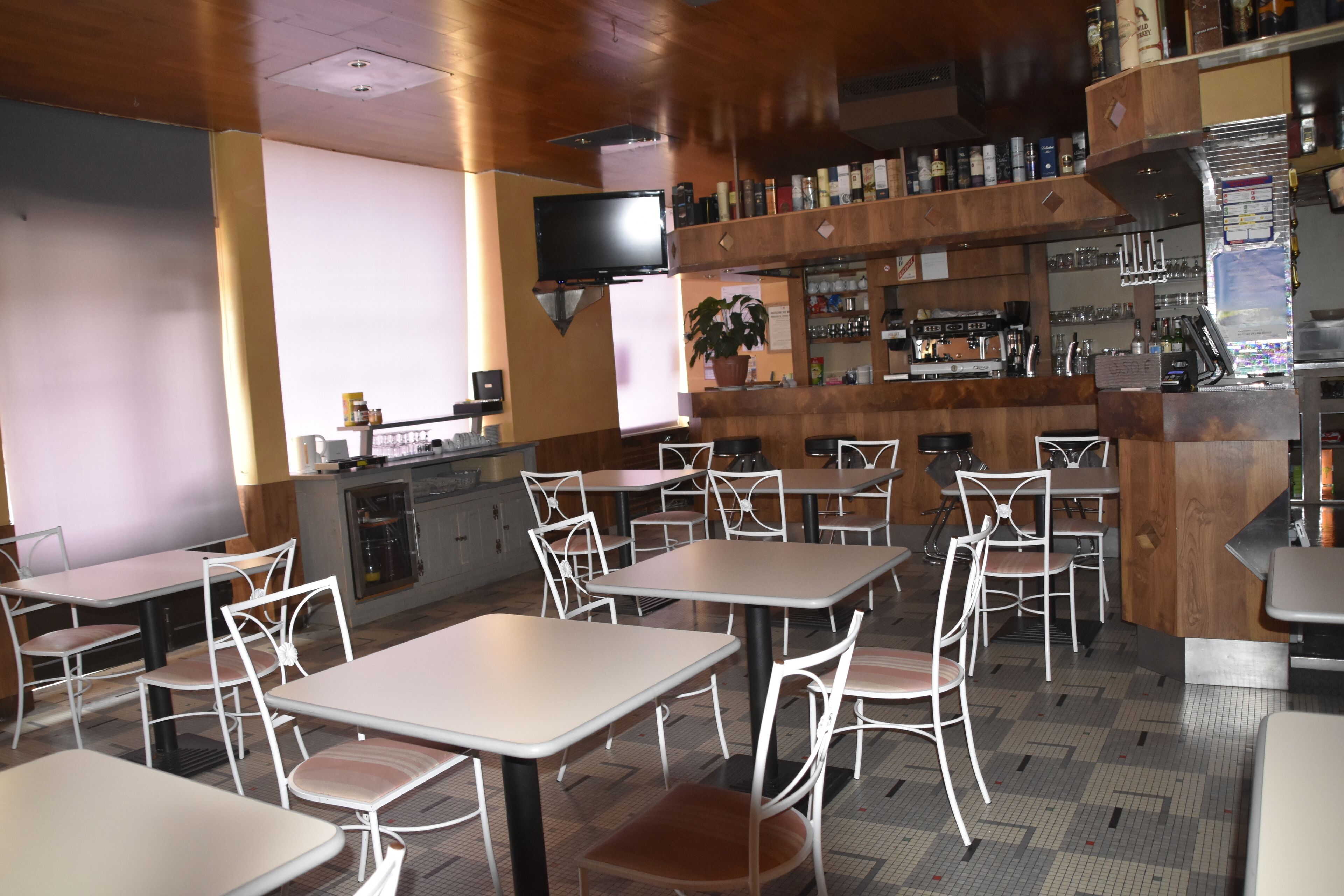 A restaurant with multiple tables and chairs, a bar area with shelves of bottles, and a television mounted on the wall.