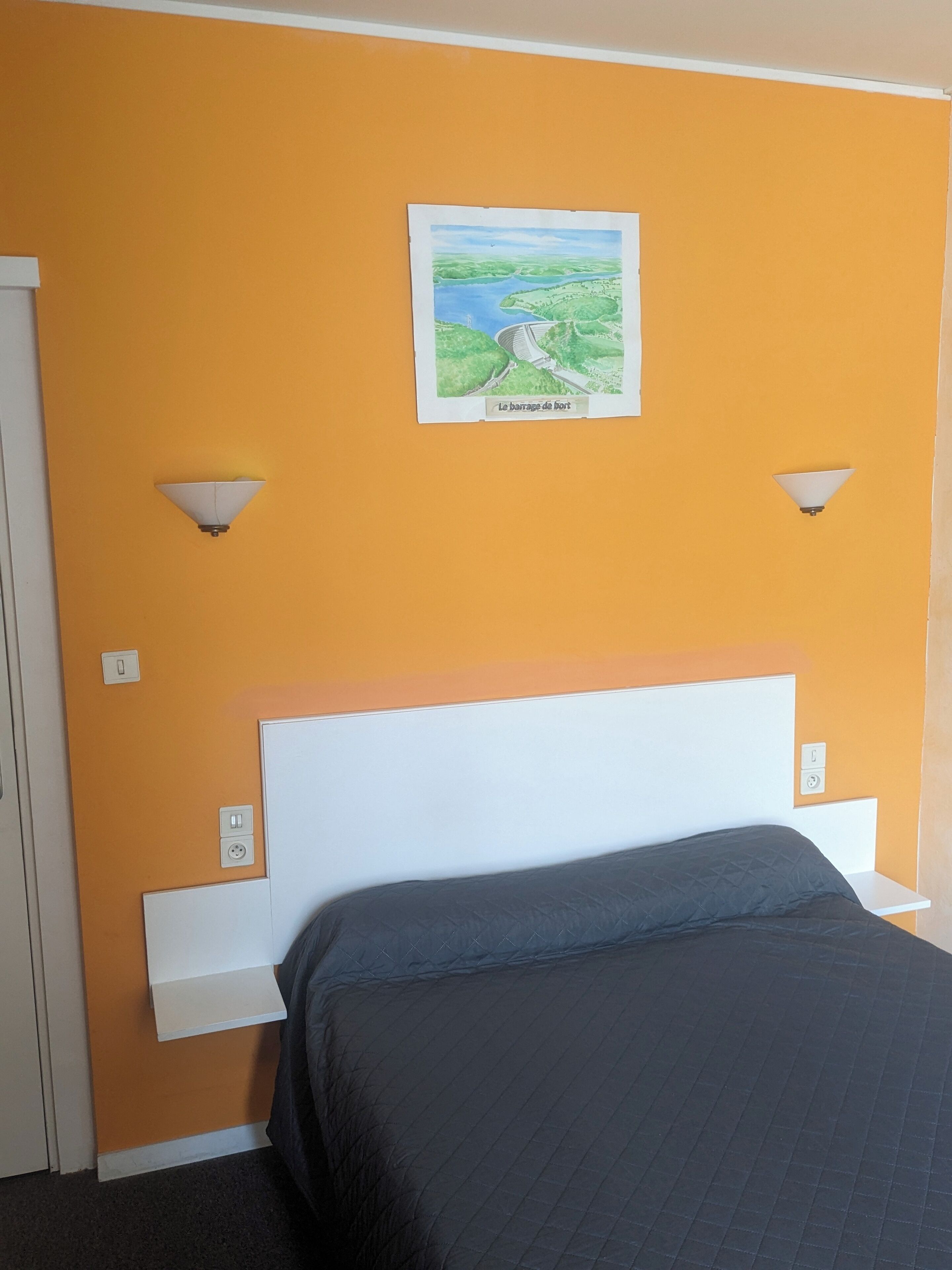 A hotel room with a bed, a white headboard, two wall-mounted light fixtures, and a framed picture of a landscape.