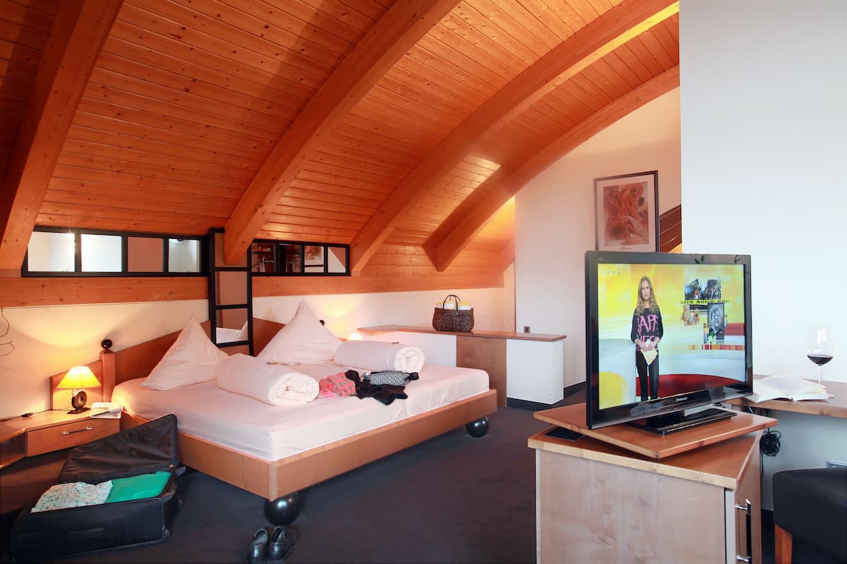 A cozy room with a wooden ceiling, a bed with pillows and a blanket, a desk with a computer and a wine glass, and a suitcase on the floor.