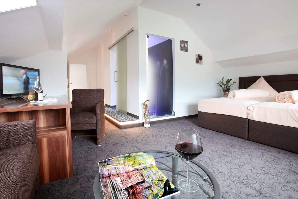 A hotel room with a desk, a chair, a bed, a glass table with a wine glass, and a small table with a magazine.