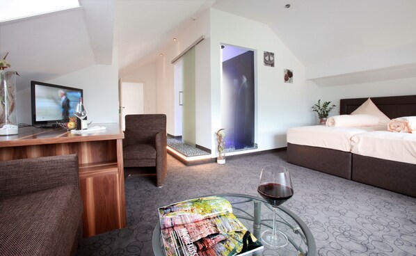 Quarto de hotel com escrivaninha, cadeira, cama, mesa de vidro com taça de vinho e uma mesinha de centro com uma revista.