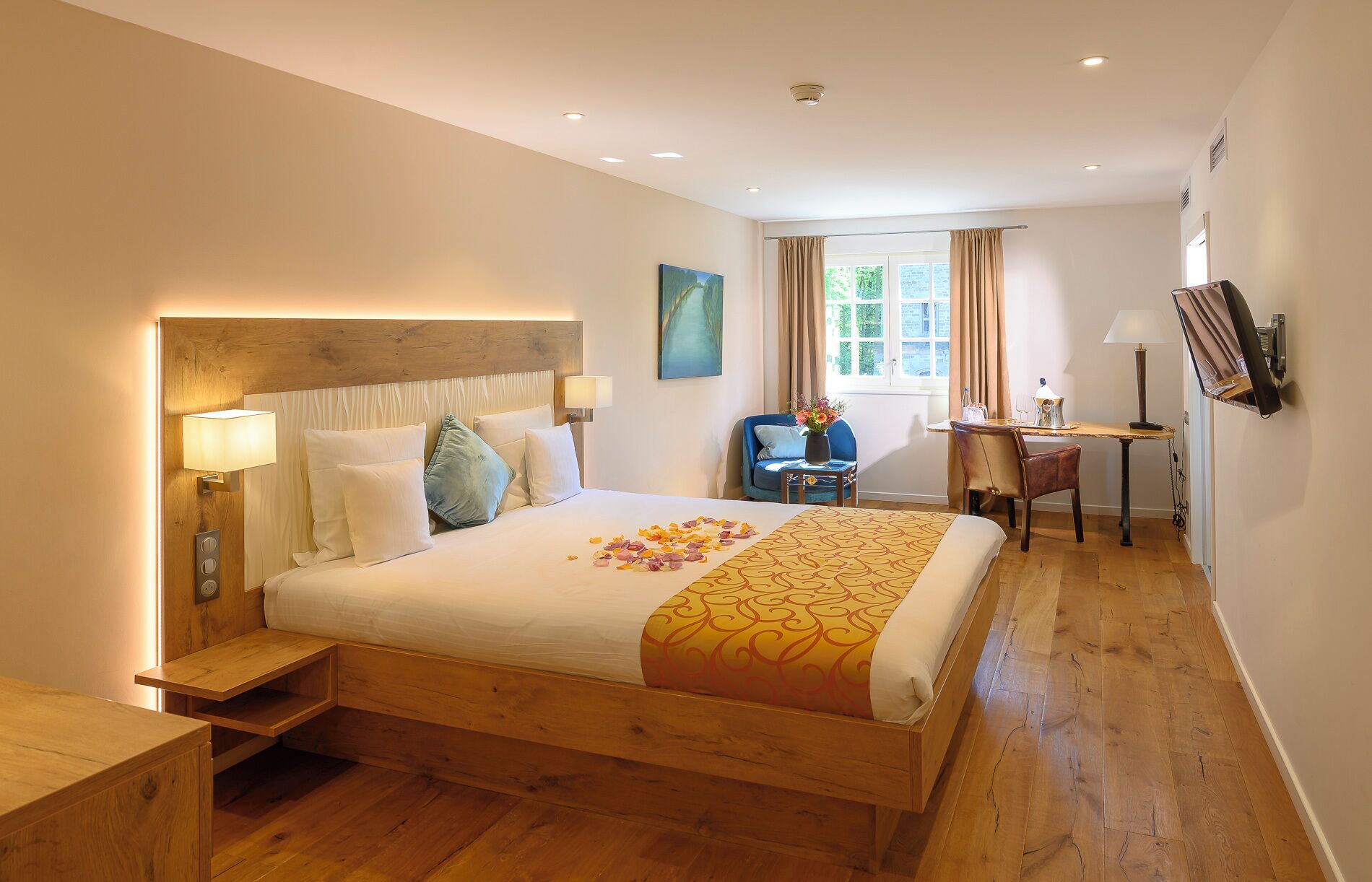 Une chambre d’hôtel avec un lit en bois, des tables de chevet, un bureau avec une chaise, une lampe et un tableau au mur.