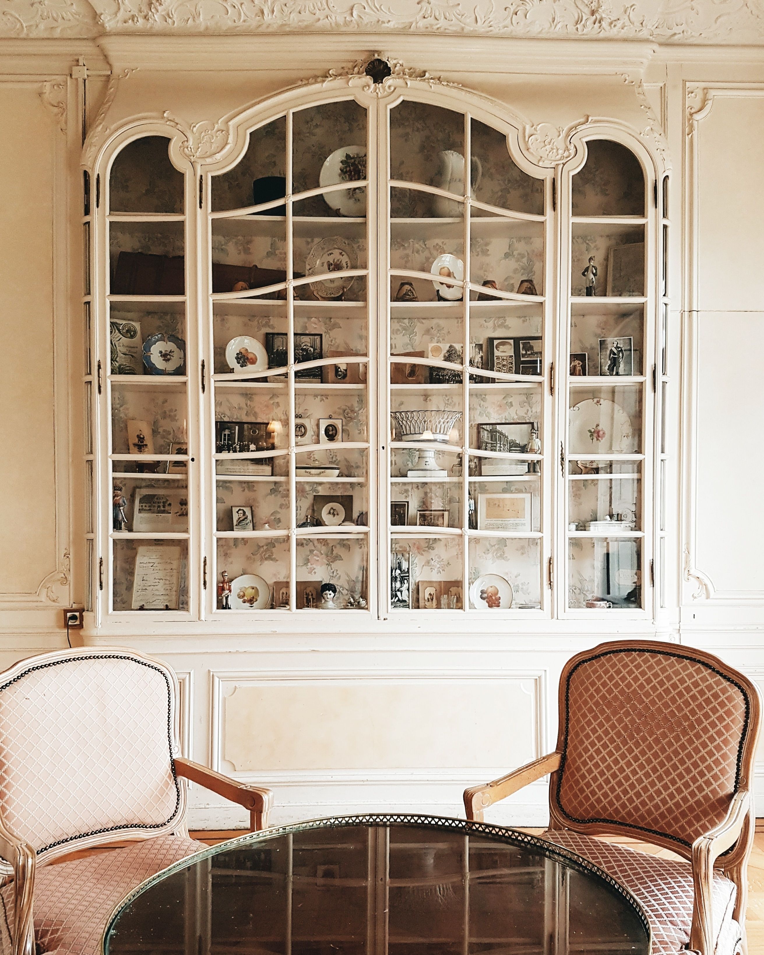 Una habitación con un amplio ventanal de arco que muestra diversos objetos decorativos. Hay una mesa redonda con dos sillas frente al ventanal.