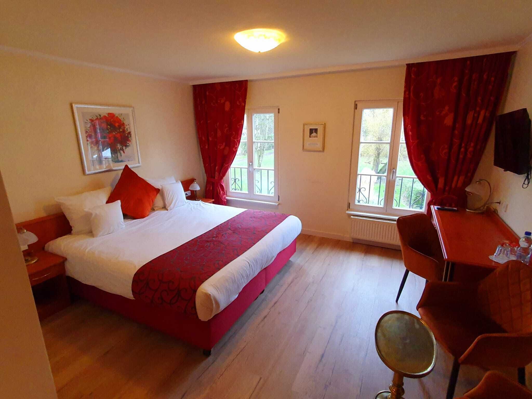 Habitación de hotel con una cama grande, dos ventanas con cortinas rojas, un escritorio con una silla y un televisor en la pared.