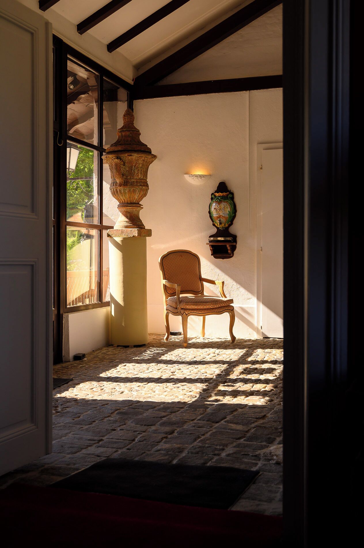 A stone-floored room with a chair, a large vase, and a wall-mounted light fixture.