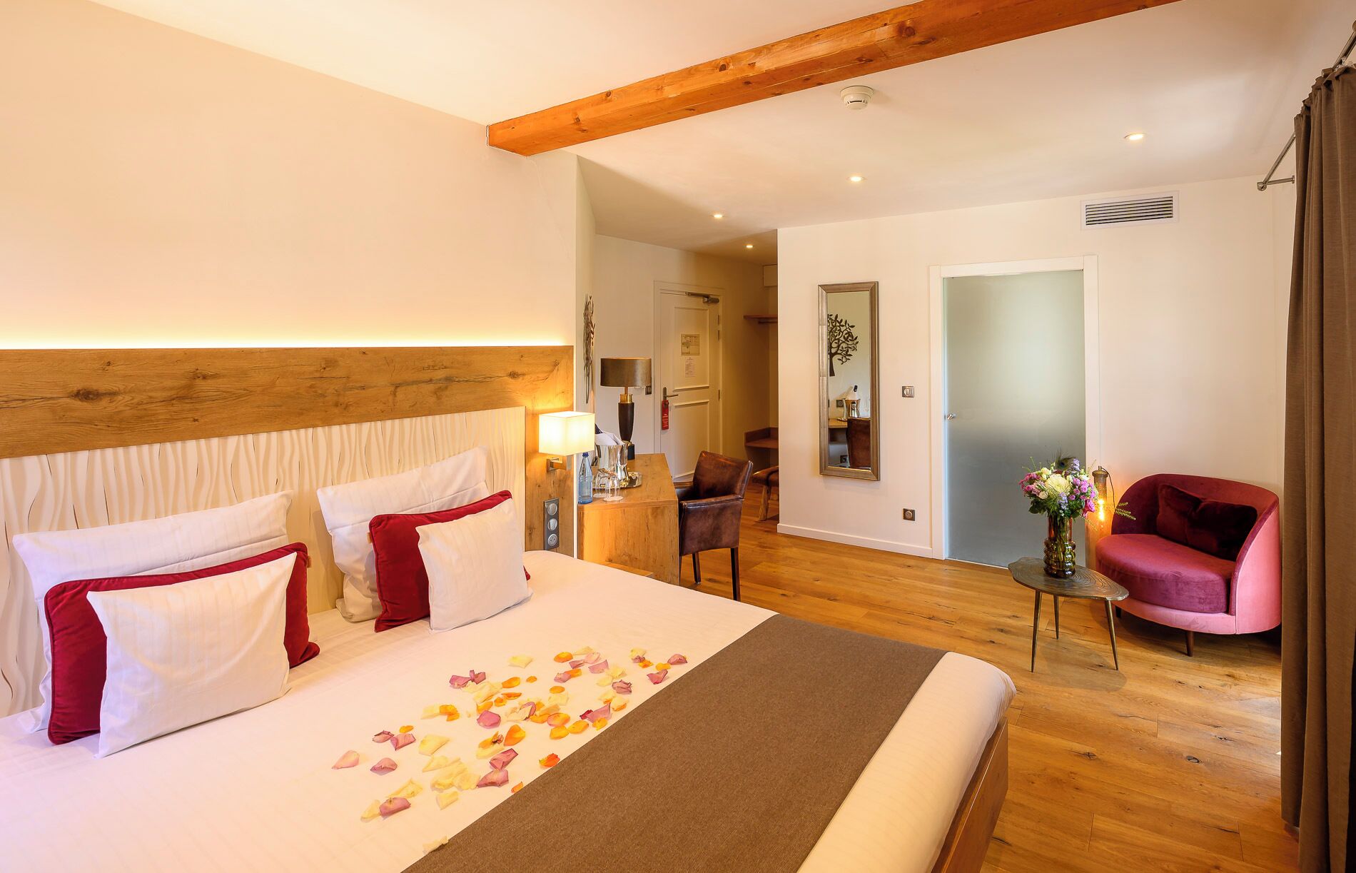 Une chambre d’hôtel avec un grand lit, une tête de lit en bois, un bureau avec une lampe, une chaise, un miroir et un canapé rose.