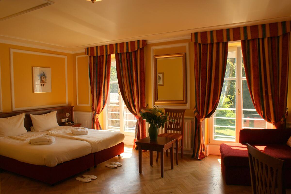 A hotel room with a bed, a table with a vase, a chair, and a window with curtains.