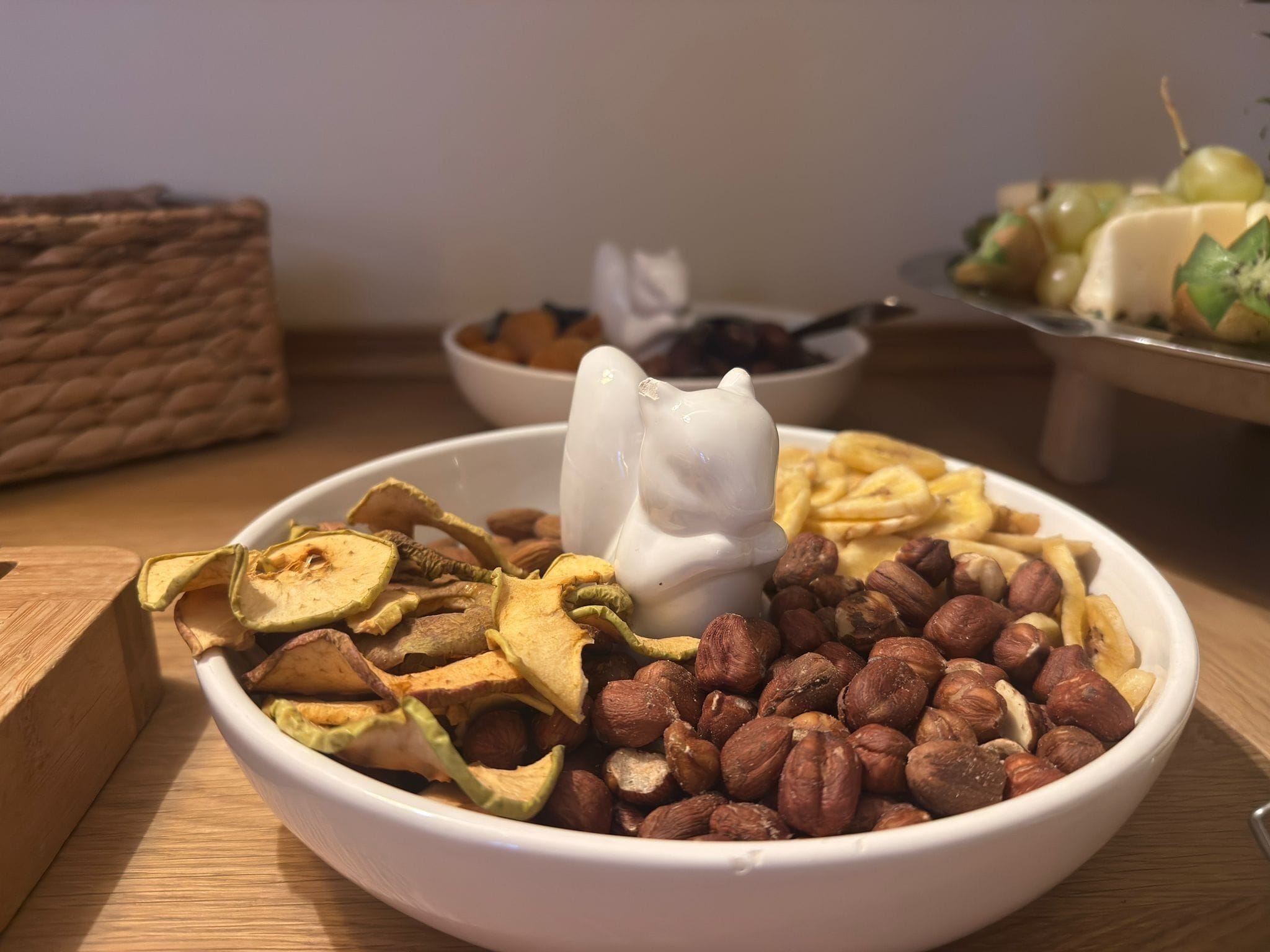 Un cuenco con frutos secos y frutas deshidratadas, junto a una cuchara con forma de gato, sobre una mesa de madera.