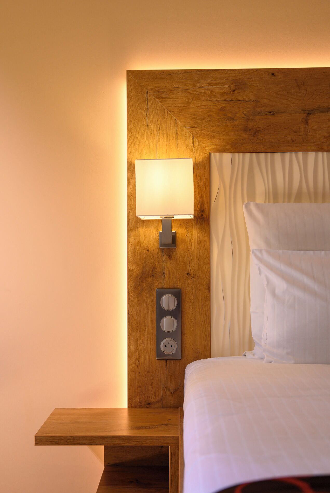 Habitación de hotel con cabecera de madera, mesita de noche, lámpara y cama con ropa blanca.