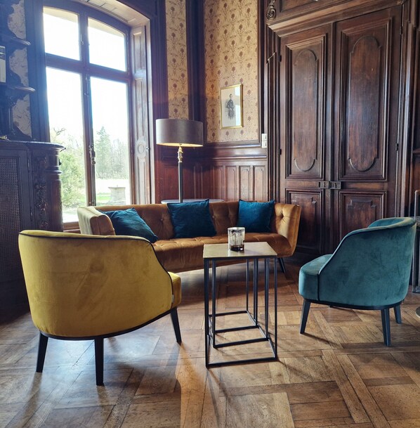 Una habitación con piso de madera, un sofá marrón con cojines azules, un sillón amarillo y un sillón verde azulado, junto a una mesa con una lámpara.