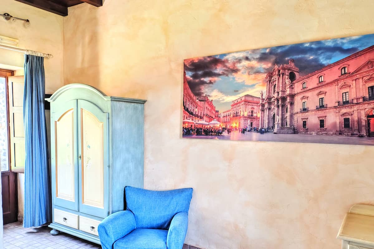 A room with a large painting of a cityscape, a blue armchair, and a vintage cabinet.