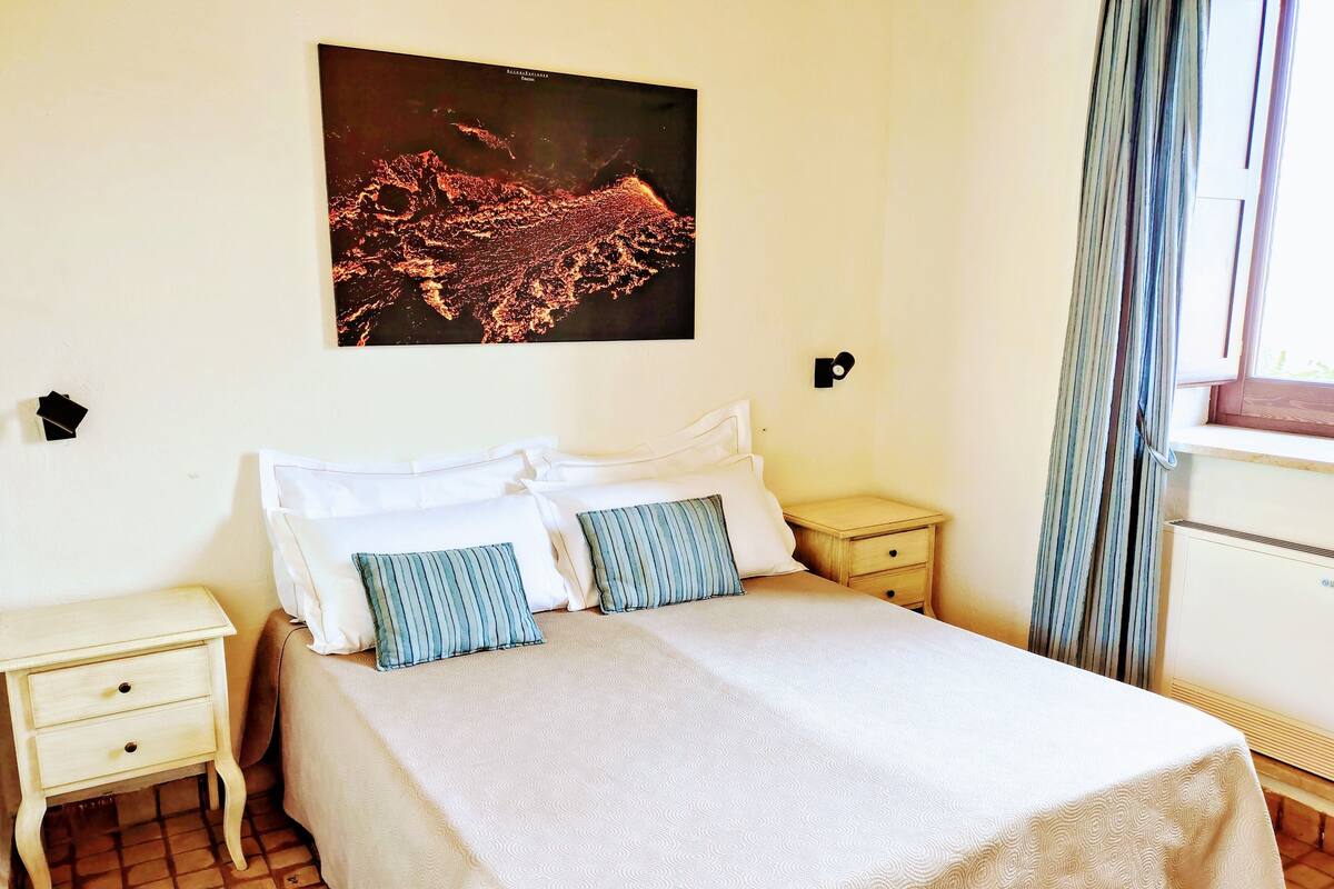 A neatly arranged bedroom with a large bed, two bedside tables, and a nightstand. A cityscape artwork is displayed above the bed.