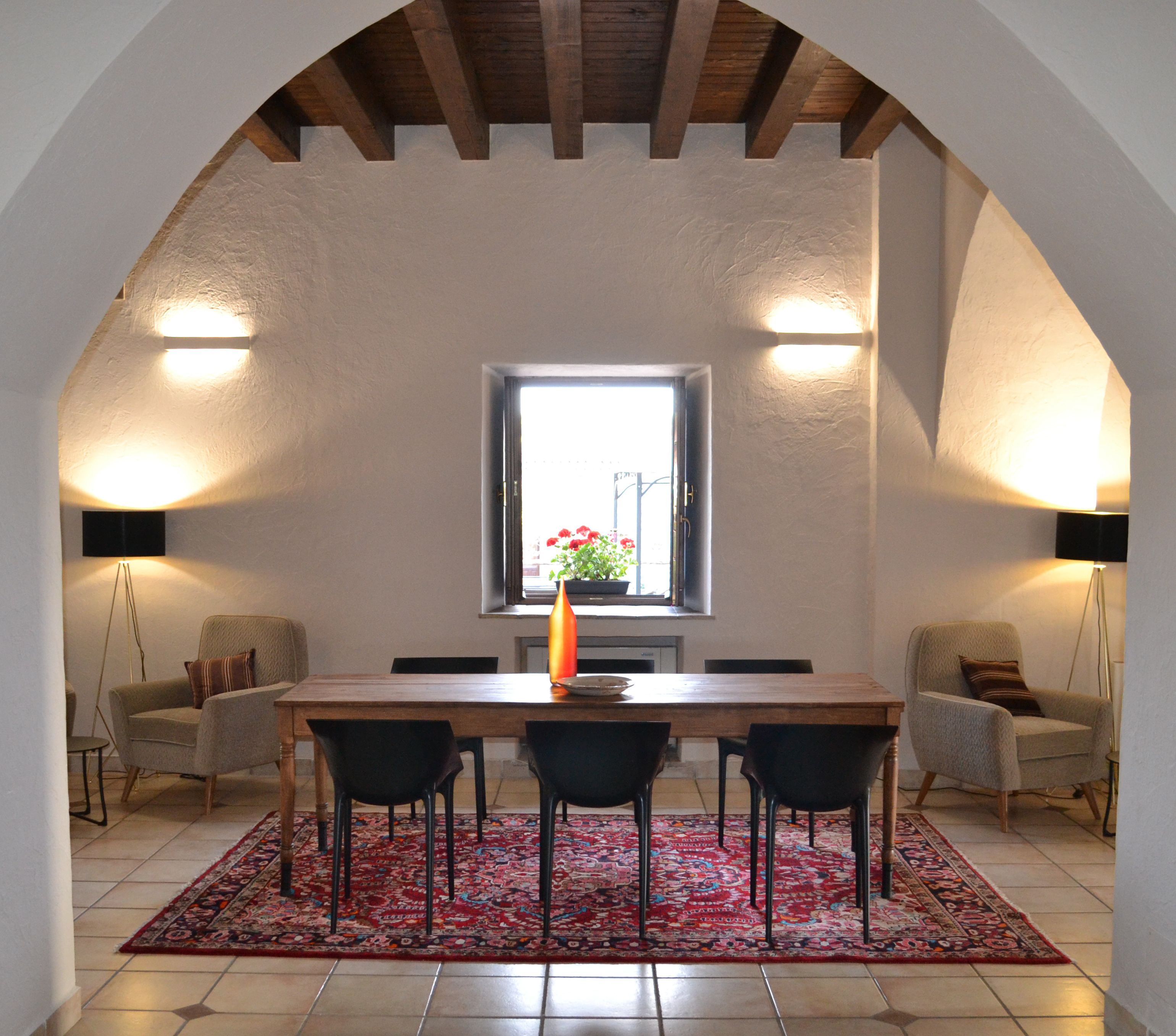A dining area with a wooden table, four chairs, a red patterned rug, and two wall-mounted lamps.