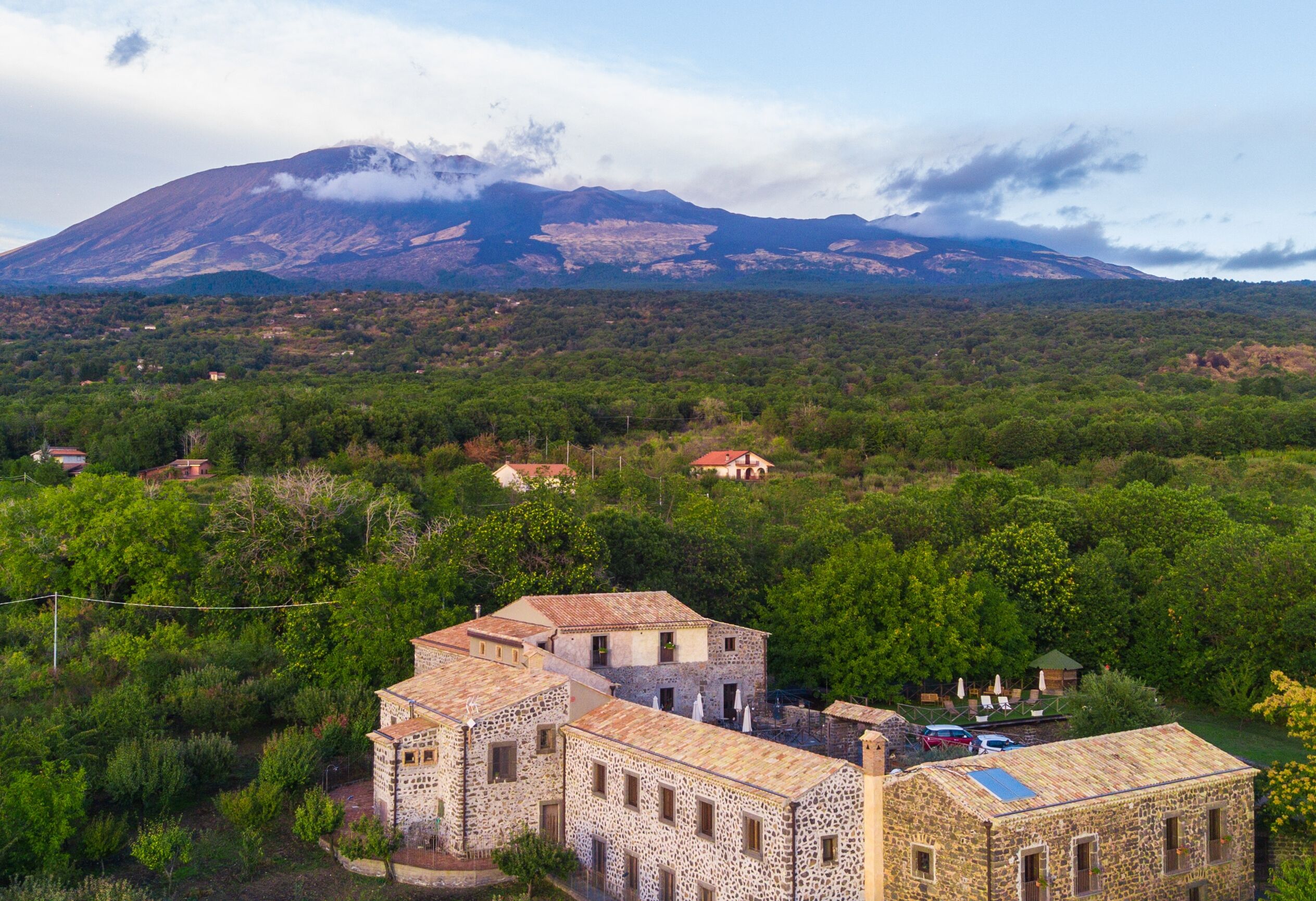 Um edifício de pedra com várias divisões, rodeado por vegetação exuberante e uma montanha ao fundo.