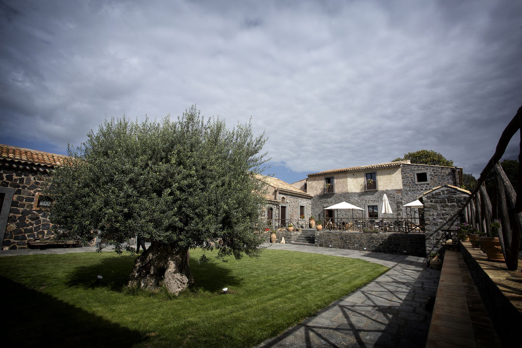 A courtyard with stone buildings, a large tree, and outdoor seating areas.