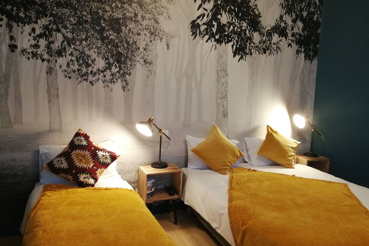 A bedroom with two beds, each with a bedside table and lamp. The room features a wall mural of trees and a blue accent wall.