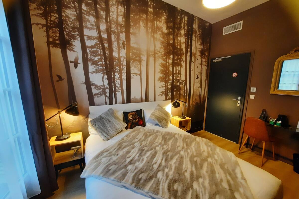 A bedroom with a bed, two bedside tables, a nightstand, a door, a chair, and a wall with a forest mural.