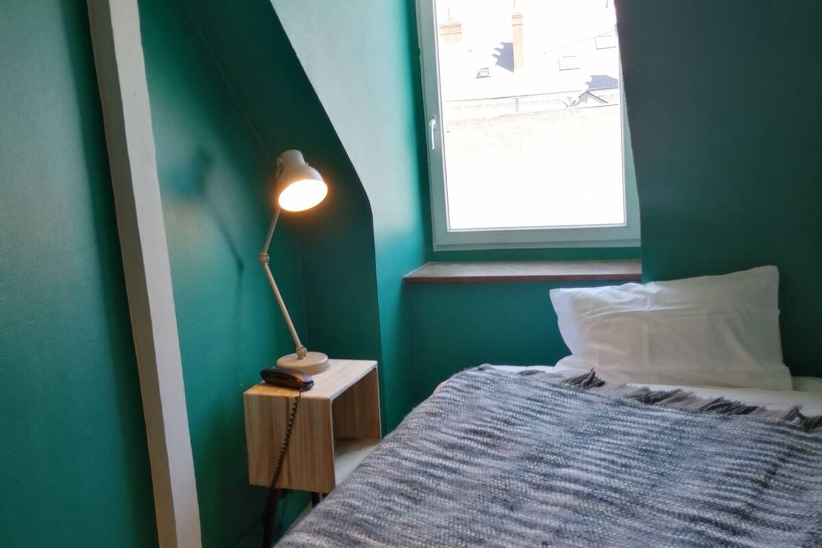 A bedroom with teal walls, a bed with a patterned blanket, a bedside lamp, and a window with a view of buildings.