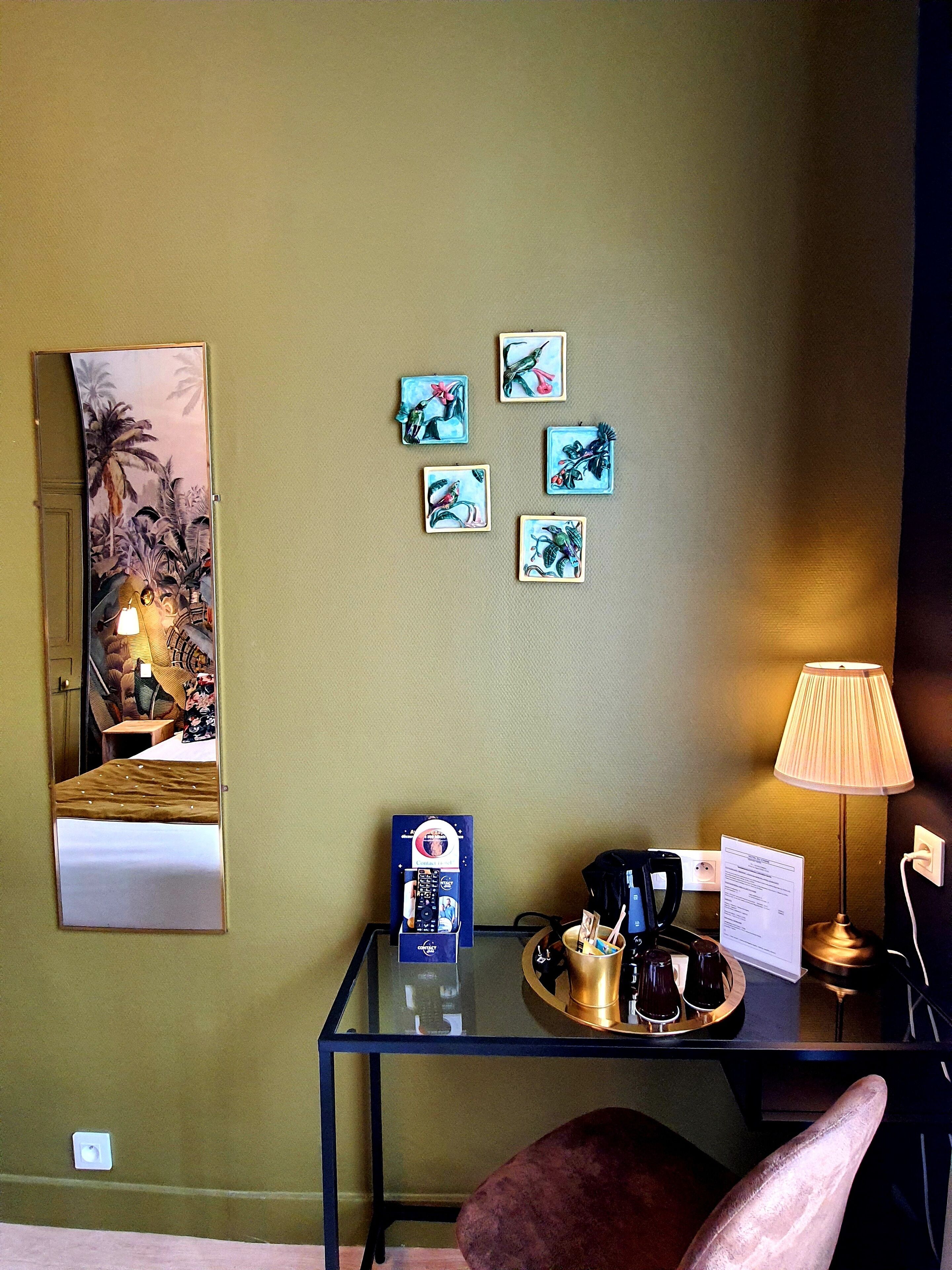A hotel room with a bed, a desk with a lamp, a mirror, and four framed pictures on the wall.