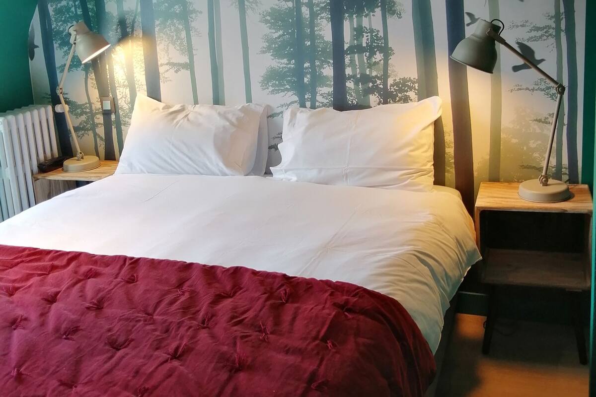 A bedroom with a large bed, two bedside tables, a red blanket, and a forest-themed wall mural.