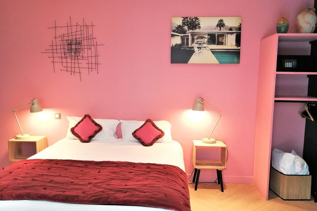 A bedroom with a bed, two bedside lamps, a red throw blanket, a pink wall, a painting of a house, and a bookshelf.