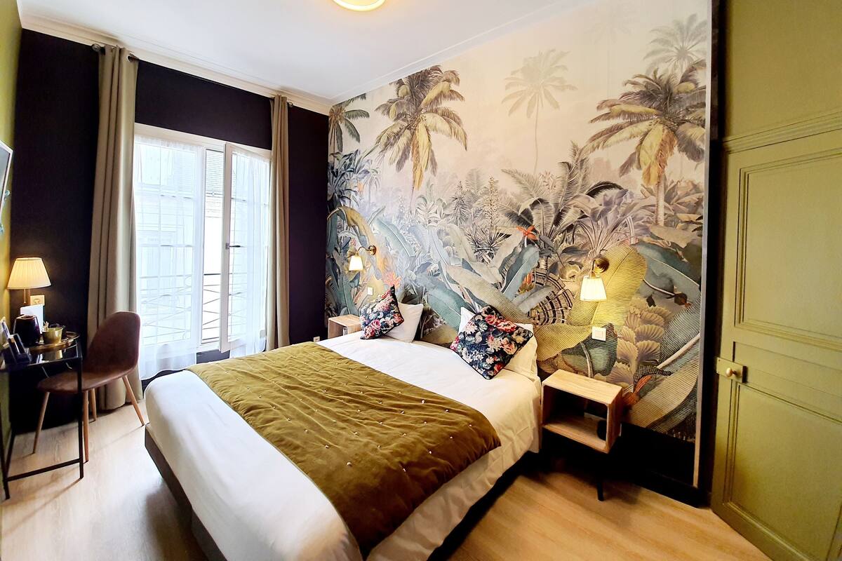 A hotel room with a large bed, a chair, a nightstand, and a wall mural of tropical plants.