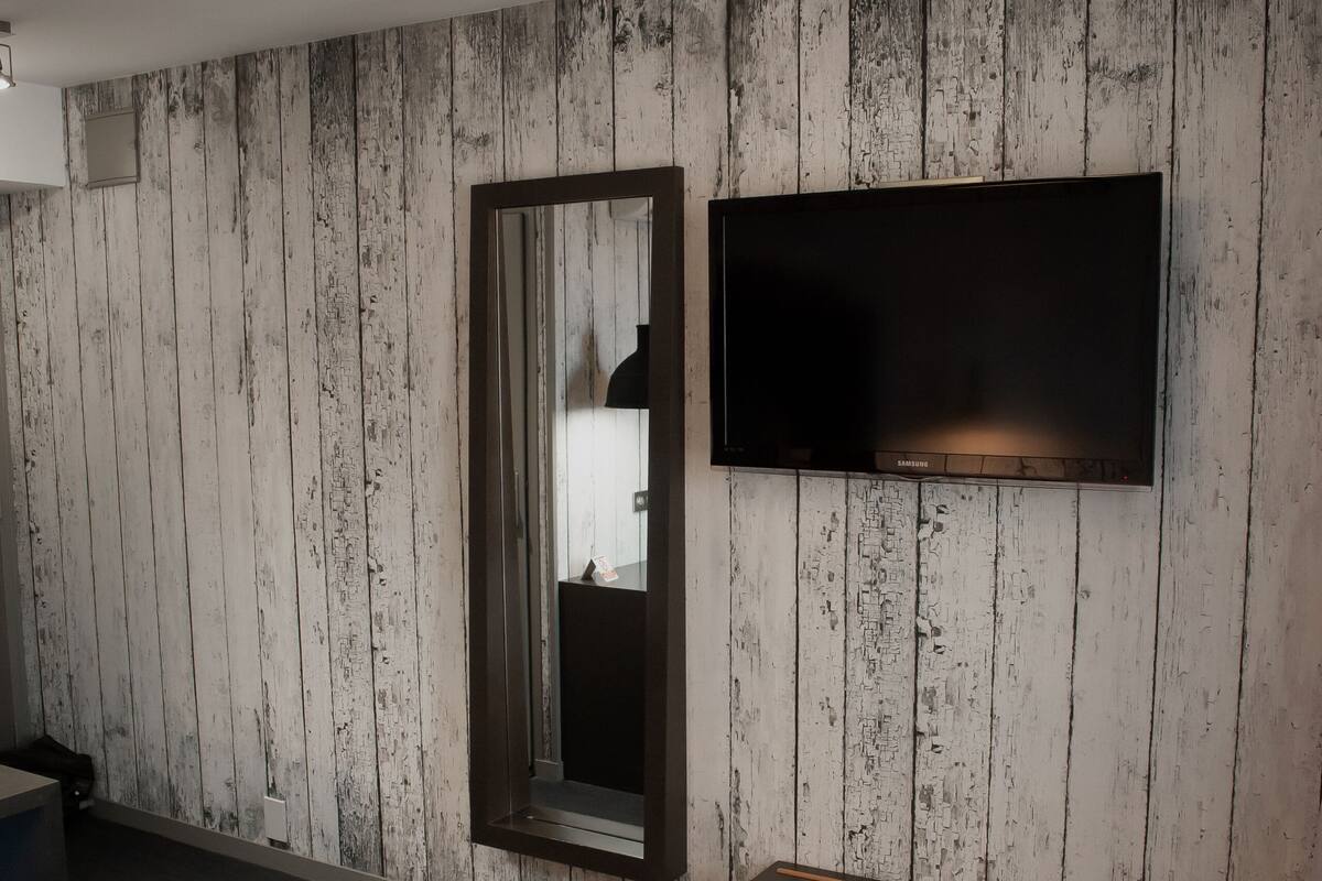 Ein modernes Hotelzimmer mit einer Holzwandverkleidung, einem flachbildfernseher über einer Holzbank und einem großen Bett mit weißer Matratze und Kissen.