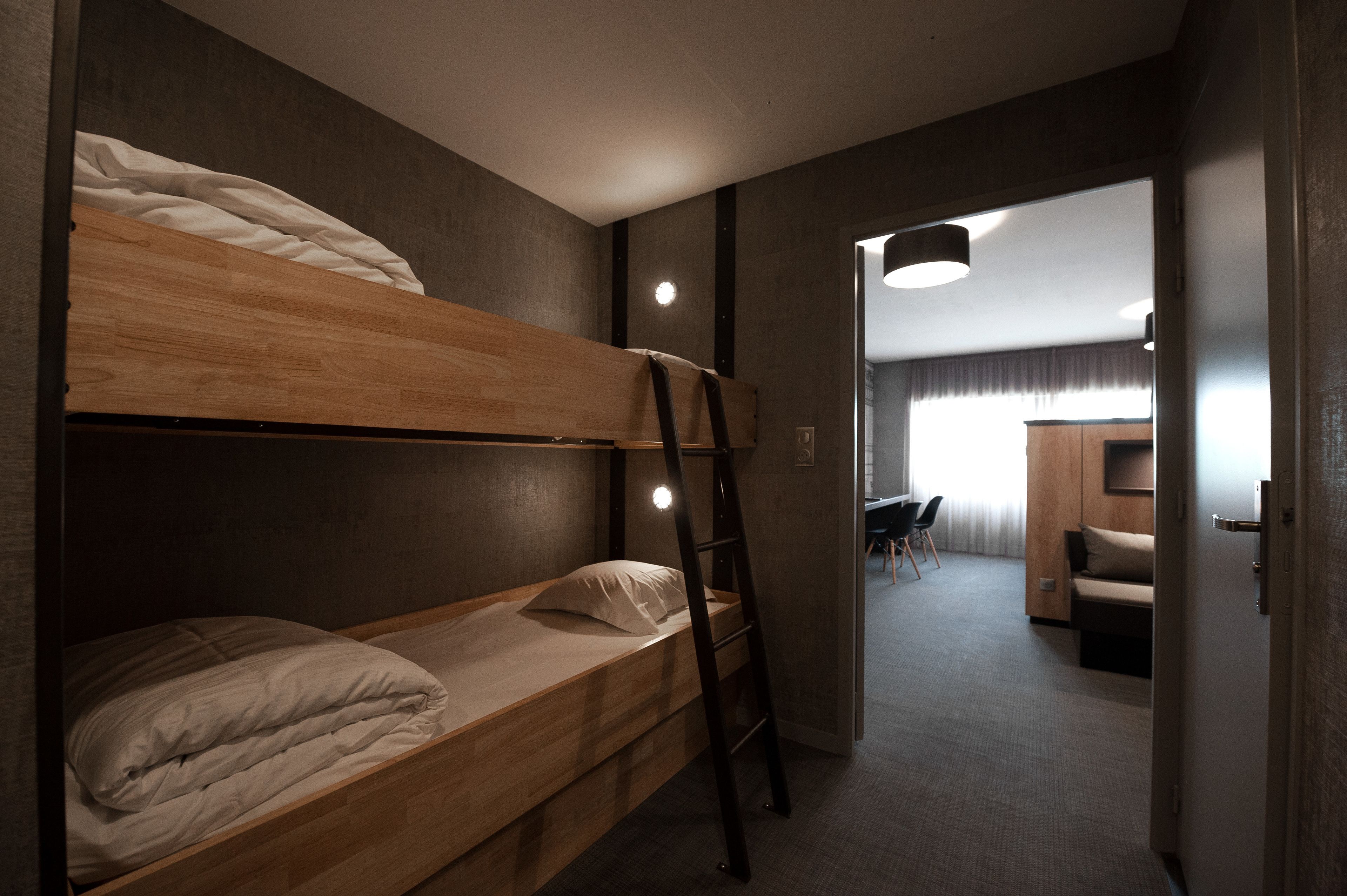 Una cama litera con escalera, un escritorio y una silla en una habitación con piso alfombrado y un acceso a otra habitación con un sofá y una lámpara.