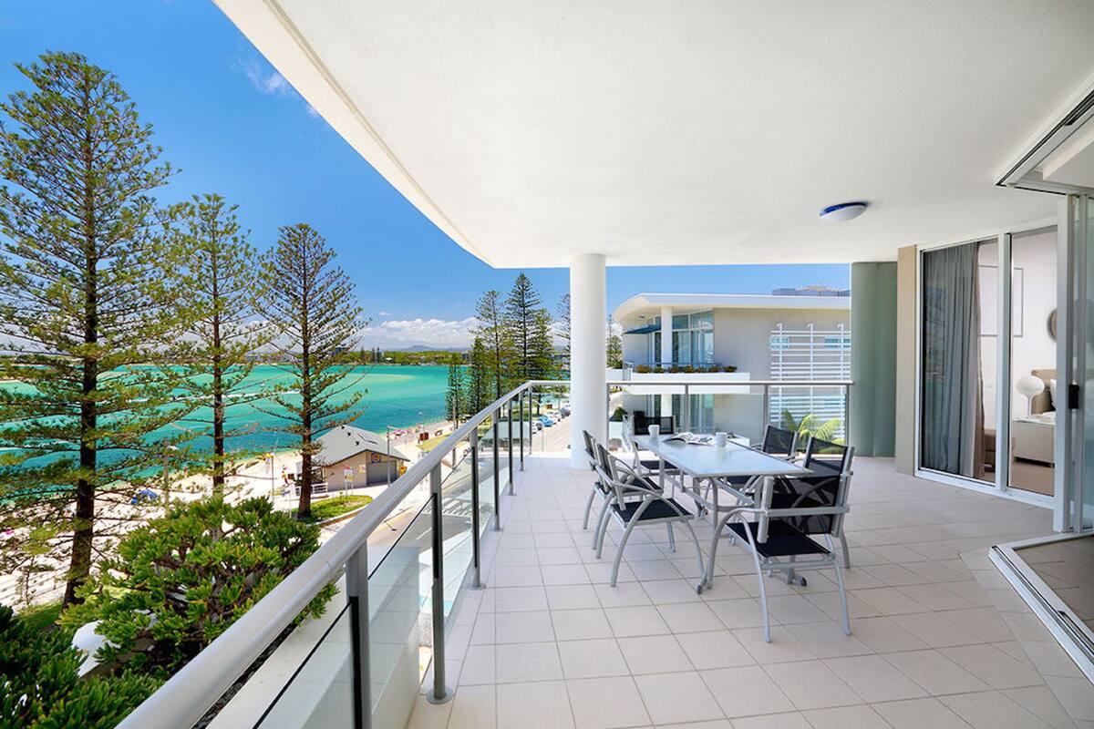 A balcony with a dining table and chairs, overlooking a beach and ocean.