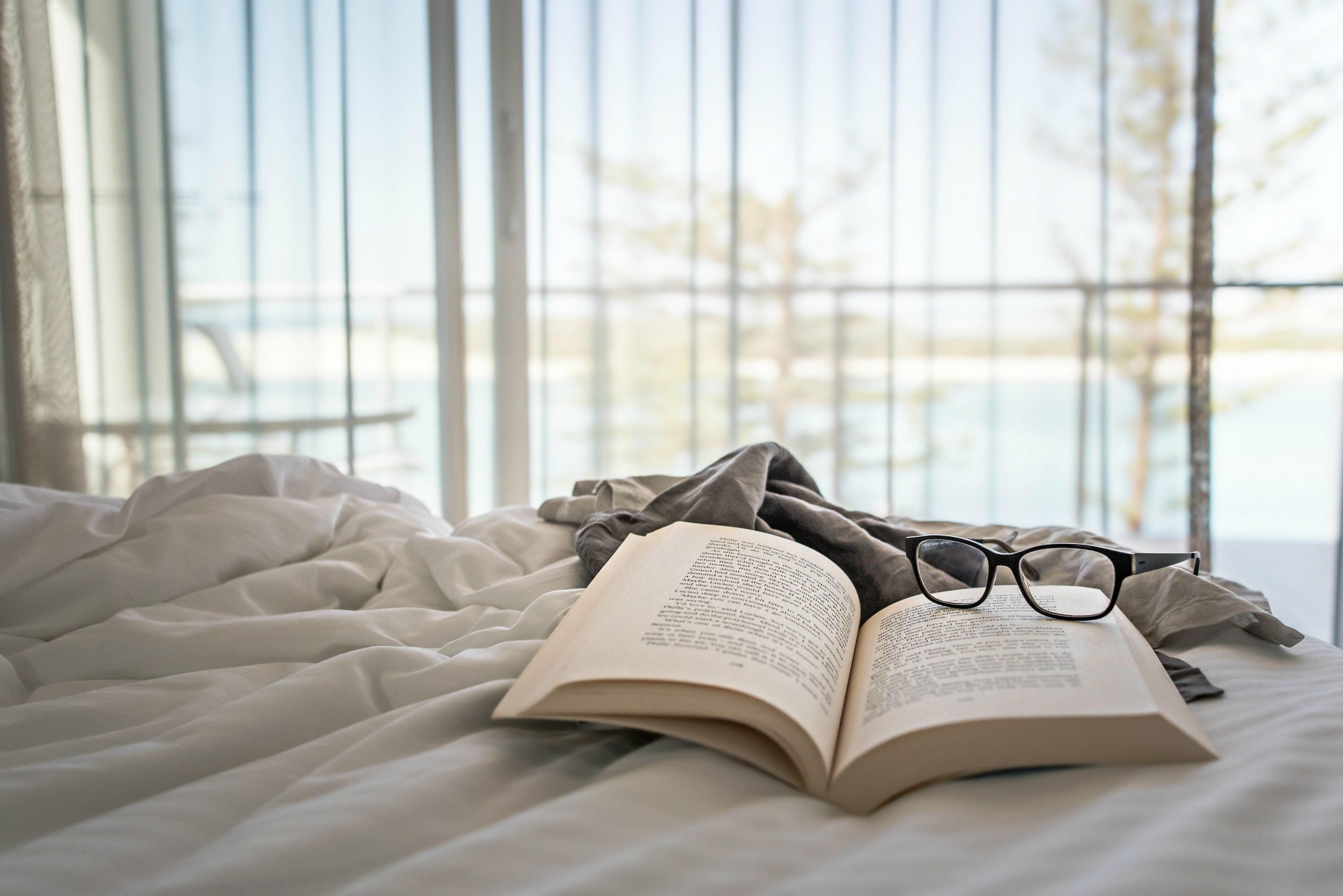 A bed with a book and glasses, a window with blinds, and a view of a body of water.