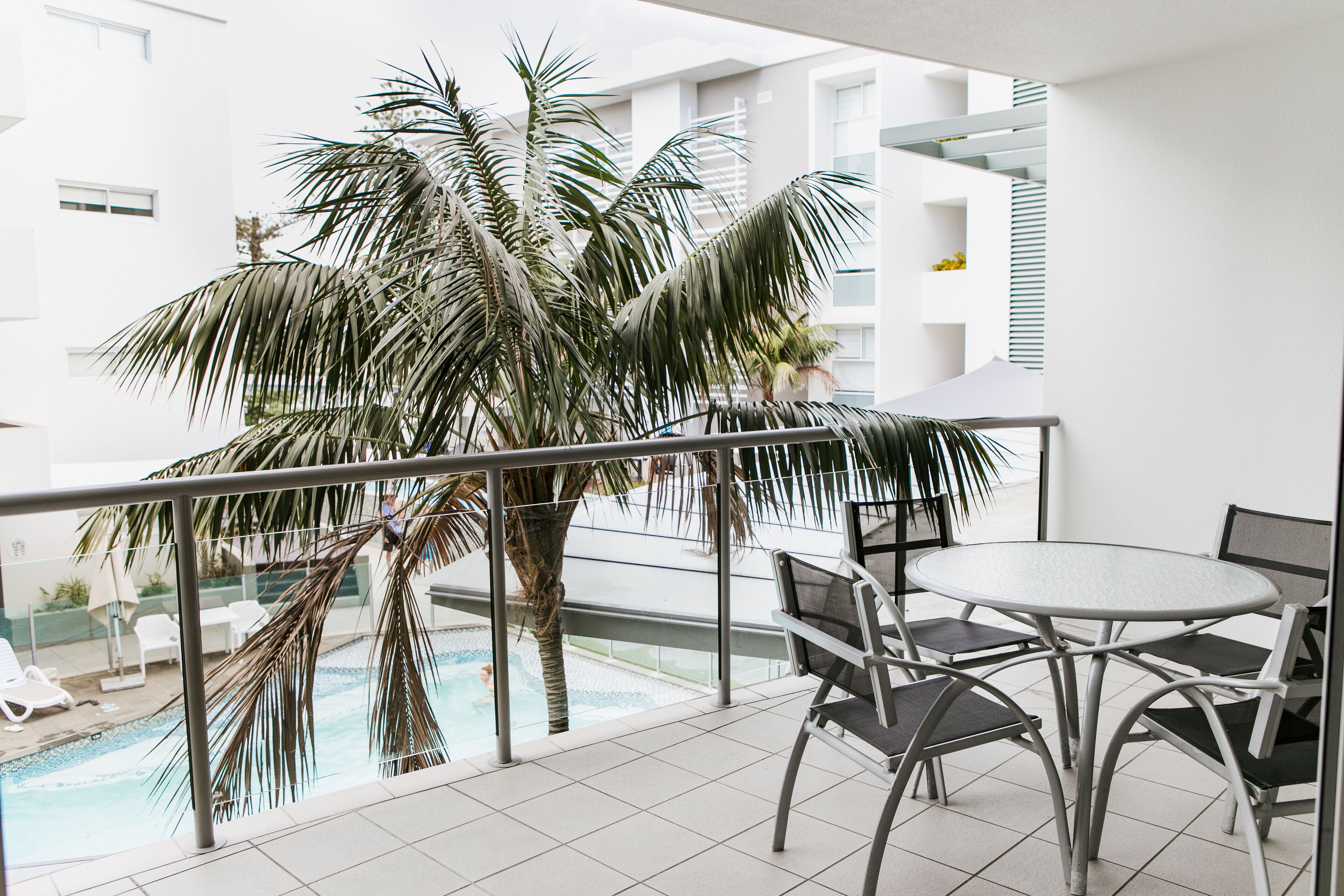A balcony with a pool, palm trees, and a table with chairs.