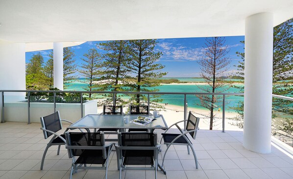A balcony with a glass table and chairs, overlooking a beach and turquoise waters.