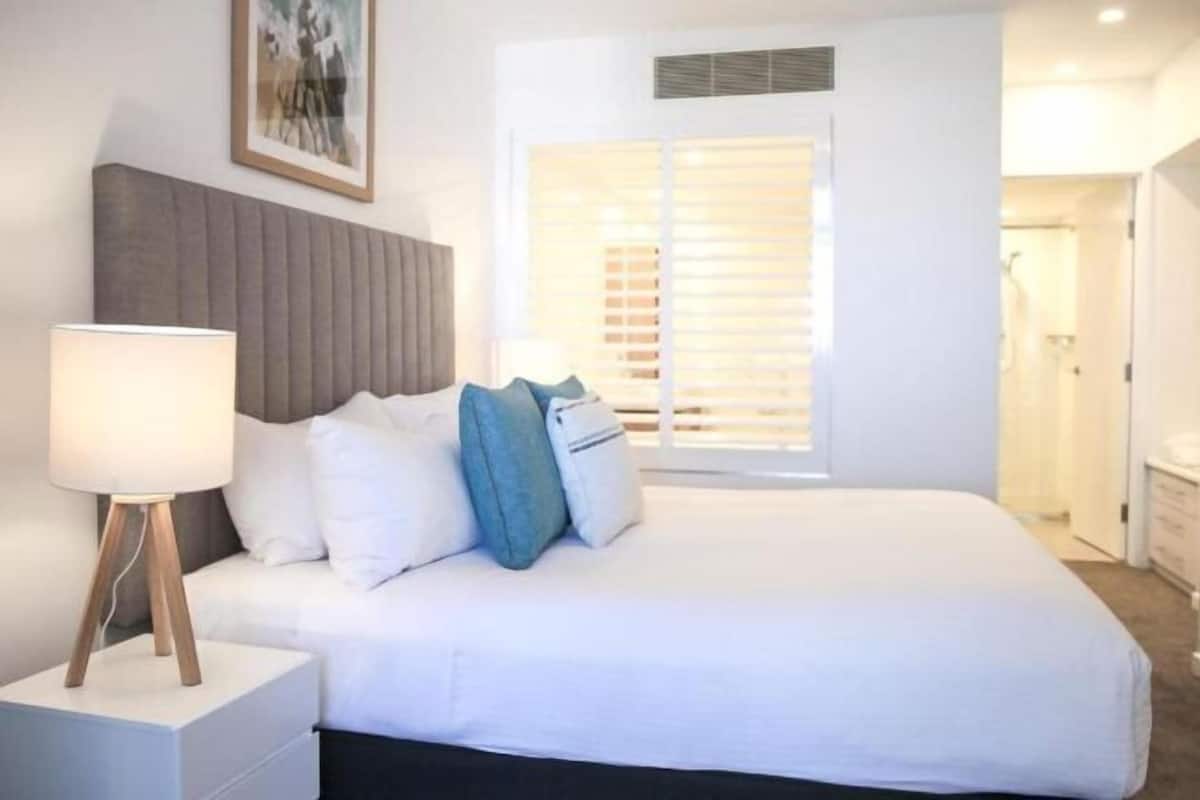 A bedroom with a bed, a bedside lamp, a window with blinds, and a framed picture on the wall.